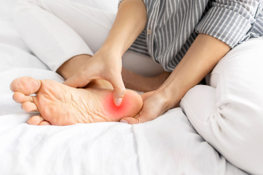 Mujer joven que siente dolor al presionar el talón y el pie mientras está sentada en la cama en casa.